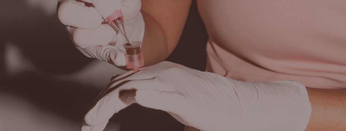 Artist dipping a PMU needle into a pigment-filled cup during a permanent makeup procedure.