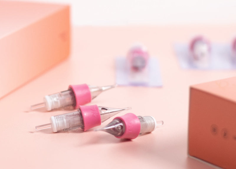 Three pink and clear tattoo needles on a light pink surface with a box in the background.